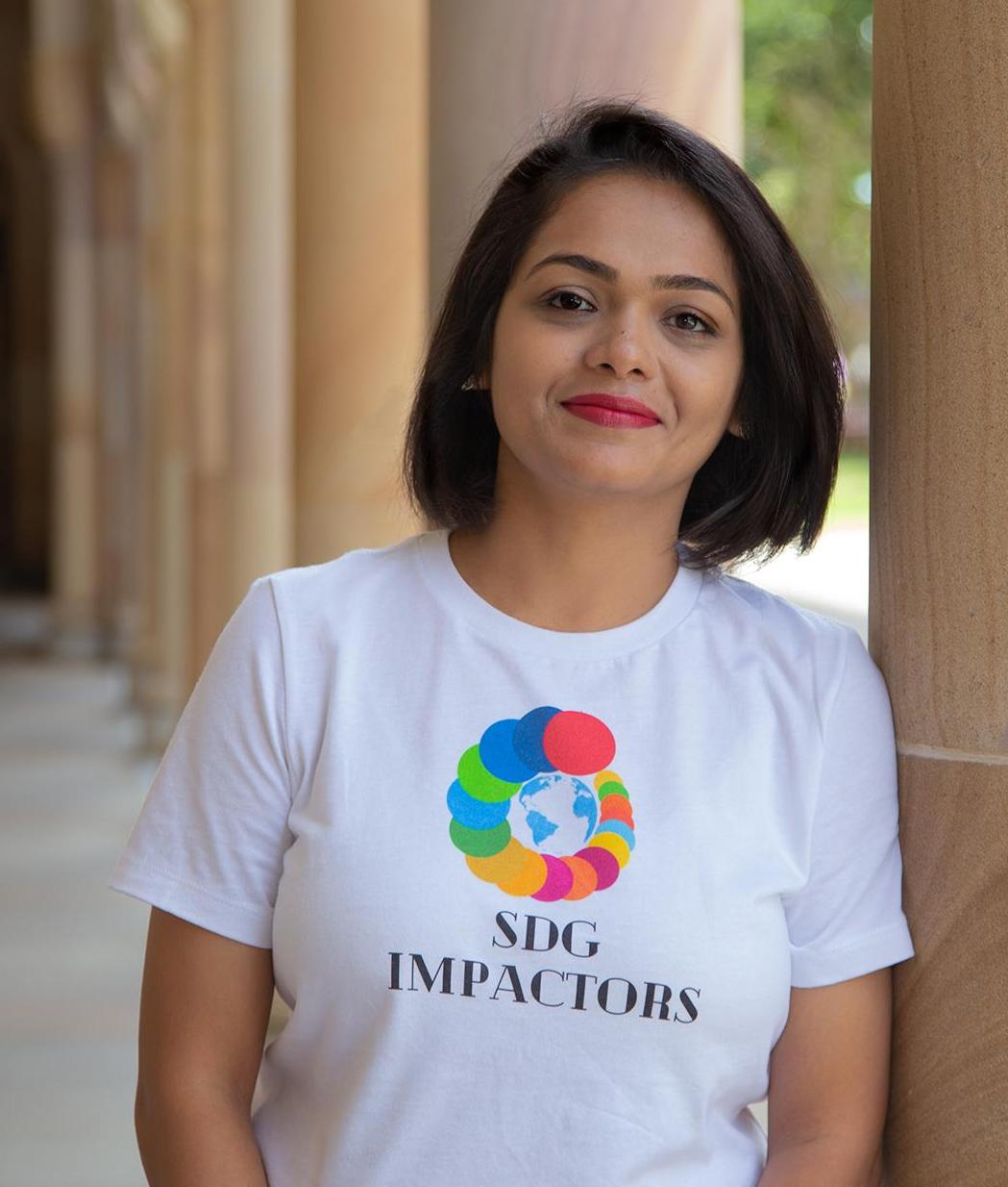 Runali smiling to camera in the University's Great Court - she's leaning against a sandstone pillar wearing a SDG Impactors' t-shirt