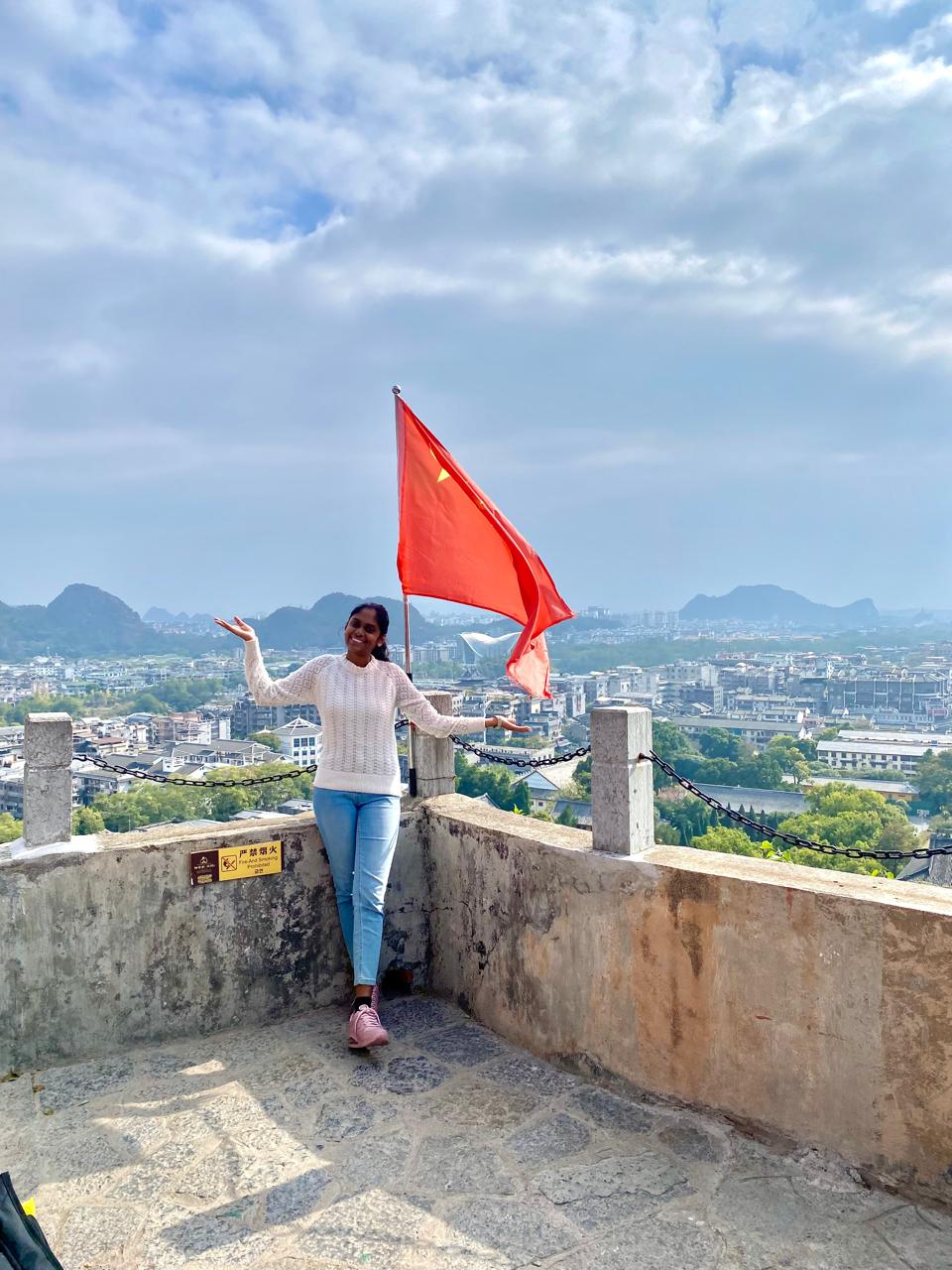 Photo of a woman in a white top and jeans standing in front of a view of a town and mountains in Shenzhen