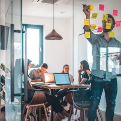 Business meeting in a glass walled office with a man putting post-it notes on the glass. You can view the room from outside the room. 