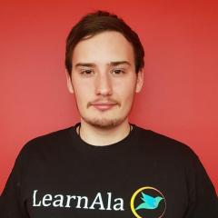 Bernardo (who has a moustache and chin beard) facing the camera with a red background behind him. He's not smiling. His t'shirt is black with the LearnAla log which has Learn and Ala (Spanish for Wing) as its image: LearnAla with winged icon.
