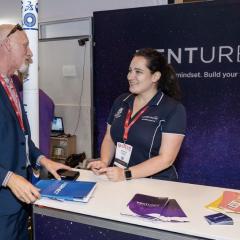 Myrthe Snoeks at QODE Brisbane Innovation Festival for UQ Ventures in 2019 with a banner behind her with UQ Ventures. She's talking to a man in his 50s. 