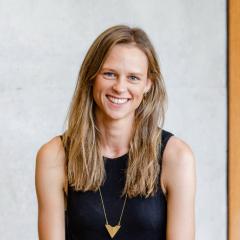 Siobhan Coster wearing sleeveless top seated inside on cement seat with wood and glass behind her. 