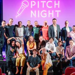 Group of people sitting and standing on stage in front out large purple screen displaying 'Pitch Night''