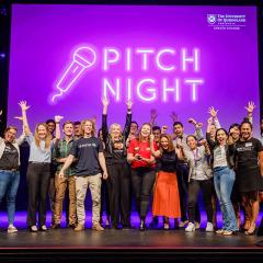 Pitch Night group shot with everyone's hands in the air - very happy vibe. Words 'PITCH NIGHT' in neon behind them. 