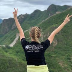 Woman in UQ tshirt in front of mountain landscape holding up peace signs